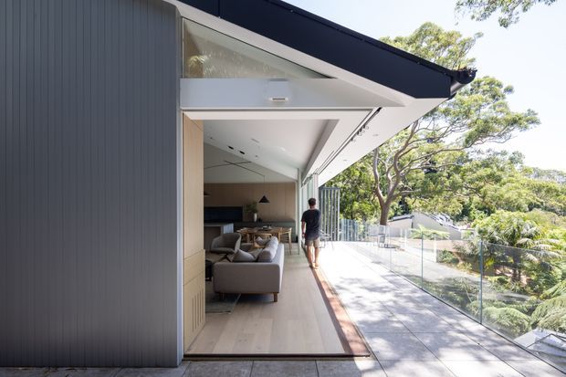 Contemporary single-storey home extension with a dark vertical-clad exterior and broad open glazed living area opening onto a long elevated terrace or balcony. The covered outdoor walkway has pale decking, glass balustrades and views through mature trees, while the interior shows a compact living room with timber built-in cabinetry and a sofa.