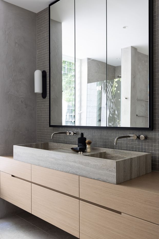 Contemporary bathroom vanity with a large black-framed mirrored cabinet, twin wall-mounted faucets, an integrated double concrete basin, light timber drawer cabinetry, dark grey small-format tiled splashback, and textured grey wall finishes. Natural light and reflected greenery add an airy, refined feel.