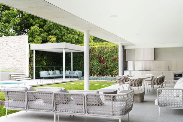 A bright, expansive contemporary outdoor living and entertaining area beneath a broad flat roof canopy. The space includes white woven lounge seating, a dining table, an outdoor kitchen with stainless-steel cabinetry, and a manicured lawn bordered by lush vertical planting. Slim white columns and a covered pavilion create a seamless connection between the interior and landscaped garden.