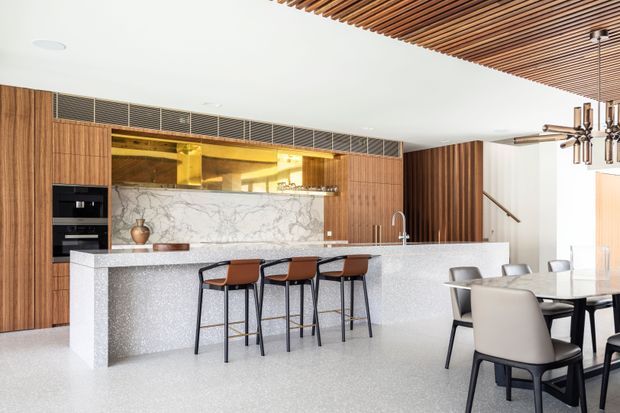 Open-plan contemporary kitchen and dining area with a large pale stone island, integrated dark appliances, warm vertical timber cabinetry, a marble-look splashback, and brass-toned display shelving. Three bar stools sit at the island, while a dining table and chairs occupy the adjacent area. A slatted timber ceiling and sculptural pendant light add architectural detail.