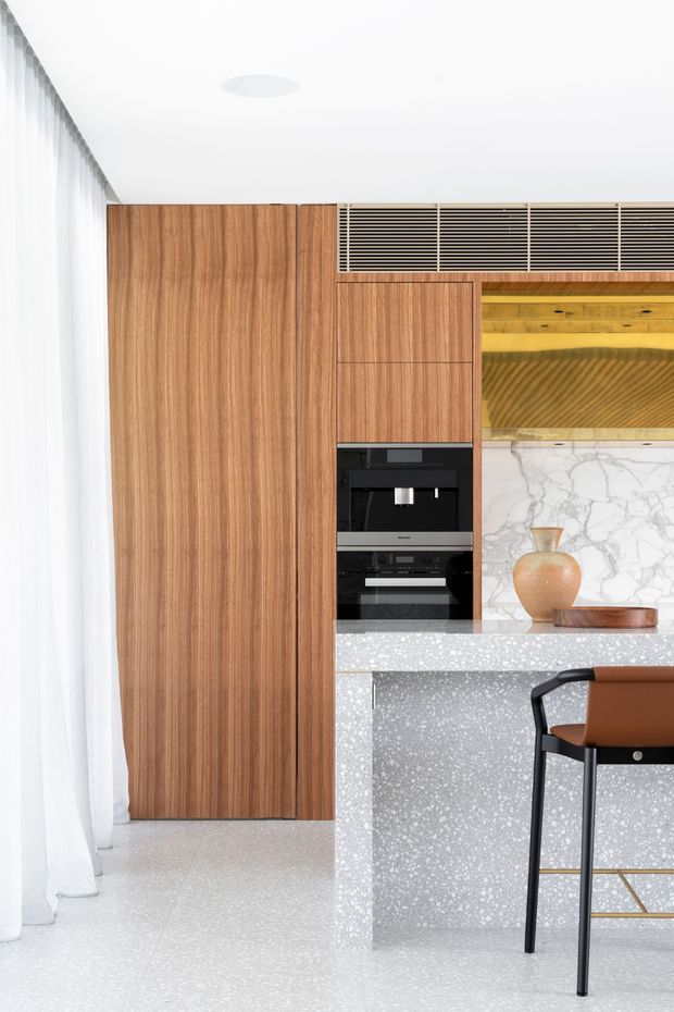 A bright, refined contemporary kitchen featuring full-height walnut-toned timber cabinetry, integrated black appliances, a large grey-and-white terrazzo island, and a dramatic white marble-look splashback. A timber-lined display niche with concealed lighting sits above the bench, alongside a black and tan bar stool. Sheer curtains and a pale terrazzo floor enhance the airy minimalist composition.