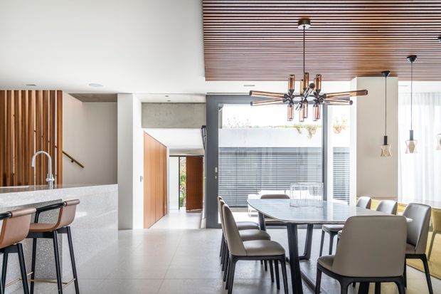 Bright contemporary open-plan dining and kitchen interior with a large dark-framed glazed opening, a long dining table surrounded by upholstered chairs, and a kitchen island with stools in the foreground. The space features pale stone surfaces, warm timber wall slats and cabinetry, a dramatic linear timber-slat ceiling, and sculptural pendant lighting.