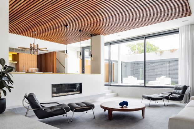 Bright contemporary living room with a sunken seating area, large black-framed windows, a linear fireplace set into a white wall, and a warm timber slatted ceiling. Black leather lounge chairs and a low round timber coffee table furnish the space, while a kitchen or bar area is visible behind a partial wall.