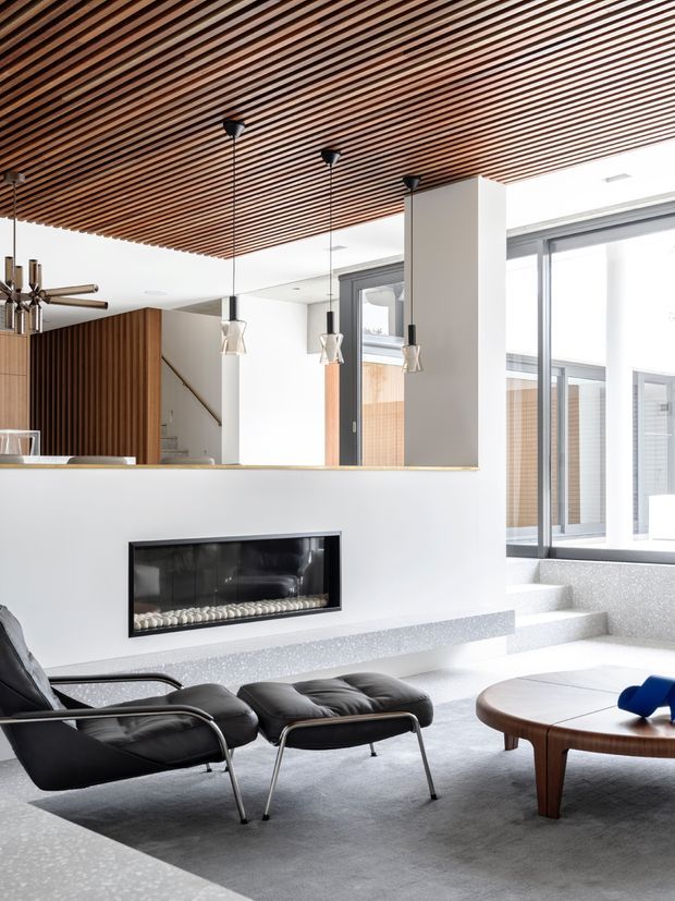 Bright contemporary living room with a linear built-in fireplace, black leather lounge chairs and ottomans, a rounded wooden coffee table, expansive glazing, and a dramatic timber slatted ceiling. A kitchen or dining area is partially visible behind the fireplace wall, with stairs and built-in timber detailing in the background.
