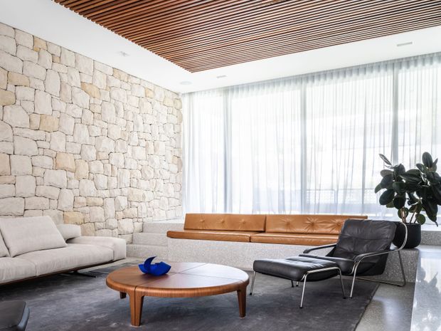Bright, refined living room with a long cognac leather built-in banquette, pale upholstered sofa, black leather lounge chair and ottoman, and a rounded mid-century timber coffee table. A rough-cut pale stone feature wall contrasts with the slatted timber ceiling, while sheer full-height curtains provide abundant natural light. A large indoor plant and minimal decor complete the space.