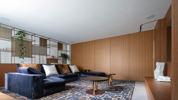 A refined contemporary living room with a large deep-blue sectional sofa, round coffee table, patterned area rug, timber slatted feature wall, and open bronze-toned decorative shelving with plants. The room has a clean white ceiling, warm timber joinery, and a sophisticated, softly layered palette.