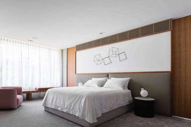 Bright contemporary bedroom with a large upholstered bed dressed in white linens, a dark charcoal headboard wall, warm vertical timber wall panelling, integrated upper ventilation grilles, sheer floor-to-ceiling curtains, and minimalist bedside furniture. A geometric wire artwork provides a subtle focal point above the bed.