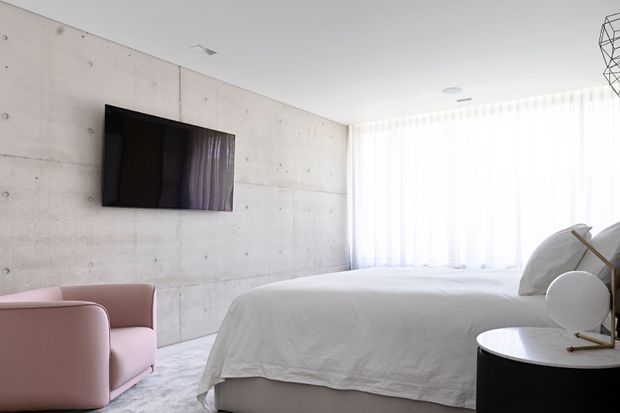 Bright minimalist bedroom with exposed pale concrete walls, a low upholstered bed dressed in white, a soft pink armchair, sheer floor-to-ceiling curtains, and a dark rounded bedside table with sculptural lighting. A wall-mounted television and restrained furnishings create a clean contemporary atmosphere.