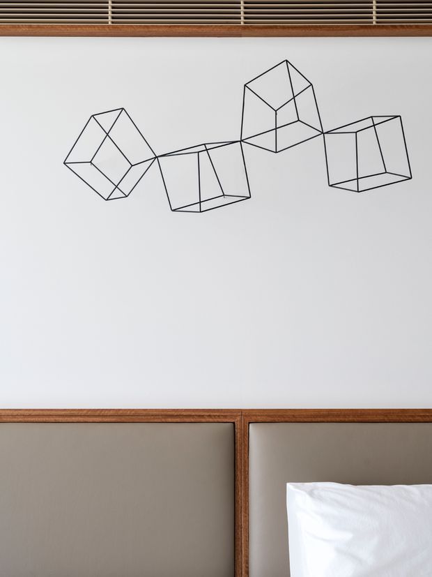 Close-up of a refined bedroom feature wall with a large white panelled surface, geometric black line artwork, and warm timber framing. Horizontal timber ventilation or slatted detailing appears above, while a taupe upholstered headboard and white bedding are visible below.