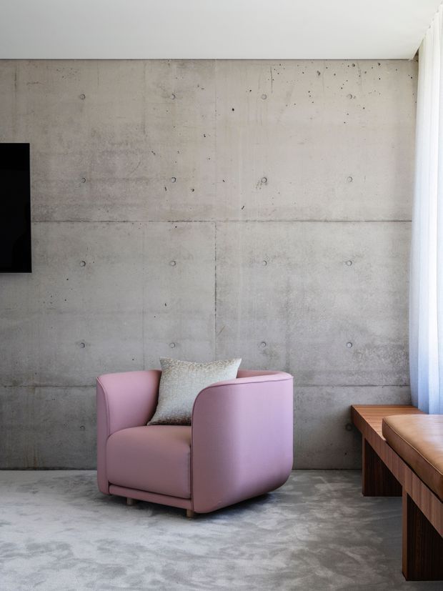 Minimalist living room corner featuring a sculptural blush-pink armchair against an exposed concrete wall, with a matching polished concrete floor, timber built-in bench or console, and sheer curtains at the window. A partial wall-mounted television is visible at left.