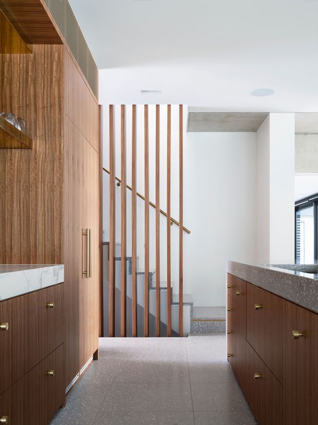 Bright contemporary interior showing a timber-lined kitchen or kitchenette adjacent to a stairway. Dark wood cabinetry with brass pulls and a speckled stone benchtop frame a terrazzo-like grey floor, while vertical timber slats screen the staircase. White walls and ceiling create a clean, refined composition.
