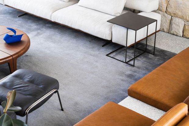 A contemporary living room viewed from above, featuring a large white sectional sofa, grey area rug, black metal nesting side tables, a black leather lounge chair, a rounded timber coffee table, and tan leather seating. A rough natural stone wall forms the background.