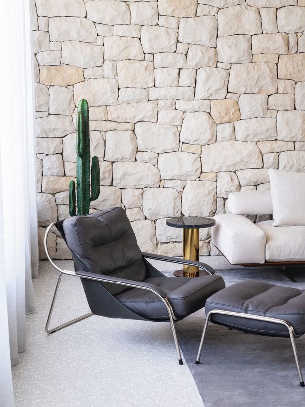A bright, refined living room featuring a dramatic irregular pale stone feature wall, a black leather lounge chair with polished metal frame and matching ottoman, a cream upholstered sofa, a small round side table with a brass-toned pedestal, a tall cactus, sheer white curtains, and a pale terrazzo-style floor. The space has a modern, minimalist aesthetic with strong mid-century furniture influences.