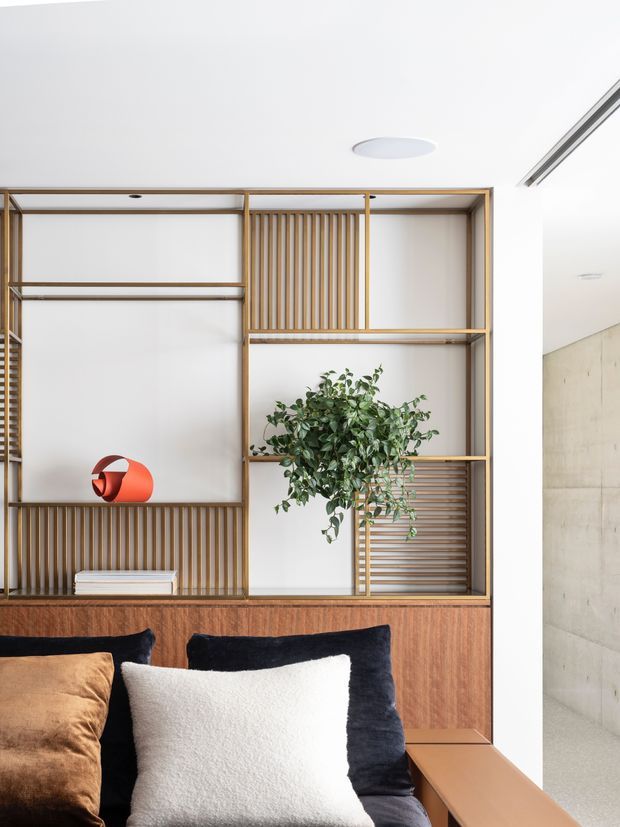 Bright, refined bedroom interior featuring a custom timber built-in shelving and cabinetry wall with open compartments, vertical timber slats, a potted plant, and a small sculptural object. The foreground shows a bed with dark navy, cream, and tan cushions. A pale concrete wall and minimalist white ceiling contribute to the clean contemporary setting.