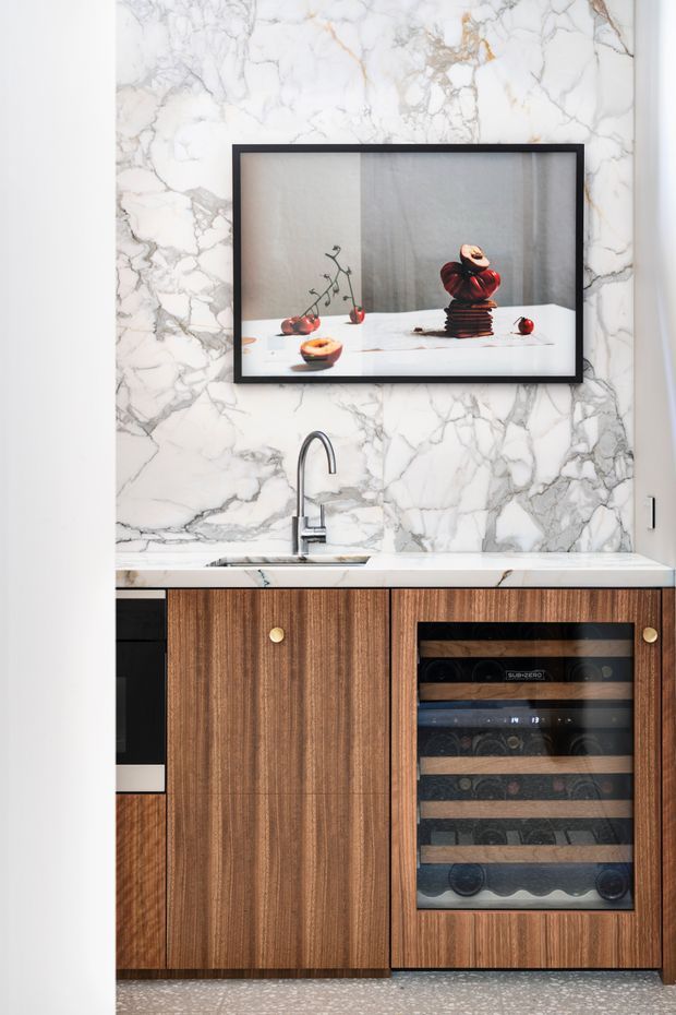 A refined kitchen or bar cabinetry vignette featuring a white marble-look wall and benchtop with prominent grey veining, a dark metal gooseneck tap, warm vertical-grain timber cabinetry, and an integrated glass-front wine fridge. A framed still-life artwork hangs above the sink, while terrazzo-style flooring is visible below.