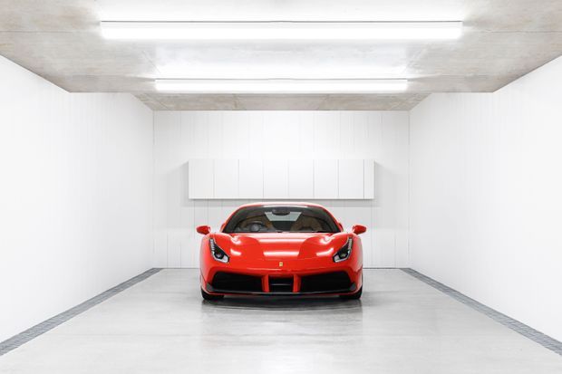 Front-facing view of a bright red sports car displayed in a minimalist enclosed garage or showroom. The space has white vertically panelled walls, a polished light concrete floor with dark perimeter strips, and a raw concrete ceiling with linear fluorescent lighting and a simple wall-mounted storage unit.