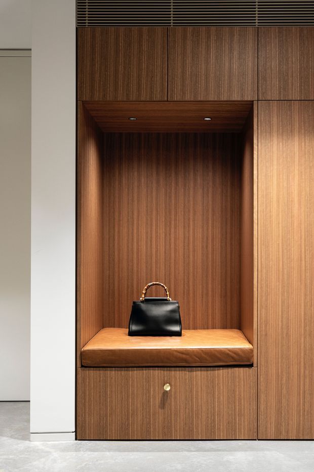 A close-up of a refined built-in timber wardrobe or entry storage unit, featuring rich vertical wood-grain cabinetry, a recessed open alcove with integrated downlights, and a tan leather cushioned bench. A black handbag sits on the bench, with a large adjoining timber cabinet door and a small brass-toned drawer pull below.