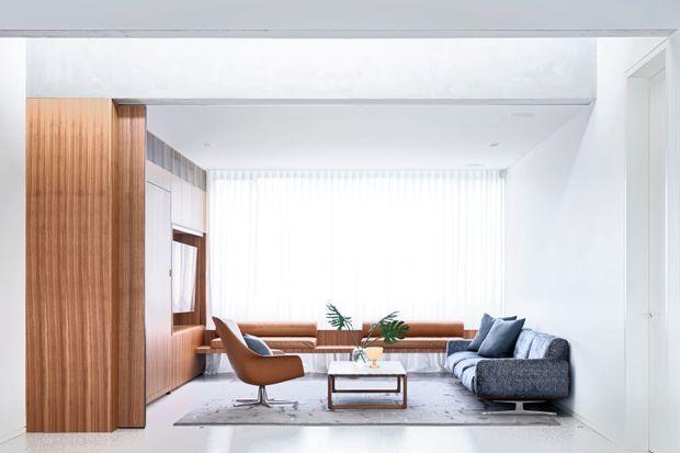 A bright, minimalist living room with white walls and ceiling, warm vertical timber wall panelling and built-in joinery, a low grey sectional sofa, brown leather lounge chair, timber-framed coffee table, and a built-in window seat beneath sheer vertical blinds. The space has a clean contemporary composition with abundant natural light and a neutral palette accented by warm wood and leather.