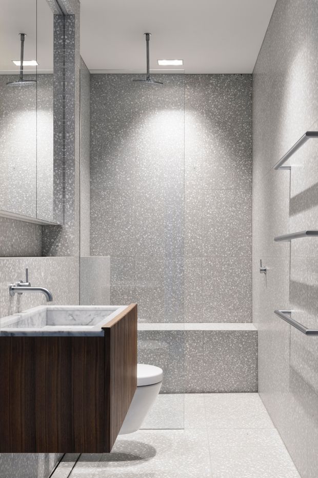 A compact contemporary bathroom with a wall-mounted dark timber vanity, white marble-look basin, chrome tapware, wall-hung toilet, large grey terrazzo-style wall and floor tiles, mirrored cabinetry, and a walk-in shower with overhead and handheld fittings. Integrated horizontal towel rails line the right wall, and bright recessed ceiling lights create a clean, refined atmosphere.