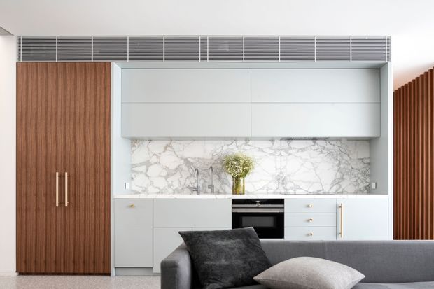 A compact contemporary kitchen viewed from an adjacent living area. The kitchen features flat-panel white cabinetry, a large white marble-look splashback with grey veining, integrated black appliances, a tall dark timber pantry or storage cabinet, and linear metal ventilation grilles above. A grey sofa with cushions occupies the foreground, while vertical timber battens appear at the right edge.