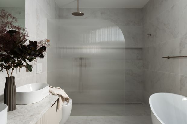 A refined contemporary bathroom with pale marble-look wall and floor finishes, a freestanding white vessel basin on a stone vanity, a partially visible freestanding bathtub, and a frameless textured-glass shower enclosure. Warm brass-toned fittings and a sculptural floral arrangement add subtle contrast. The room is softly lit and serene, with a rounded shower ceiling detail.