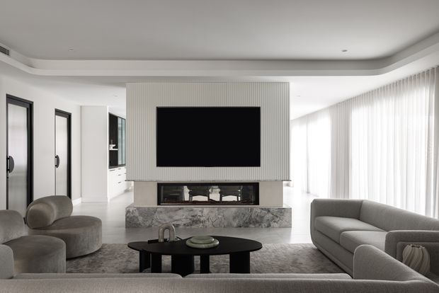 A bright, refined contemporary living room with a large sectional sofa, rounded lounge chairs, black oval coffee table, marble fireplace surround, vertically ribbed feature wall with wall-mounted television, and expansive sheer-curtained glazing. The room uses a restrained palette of white, grey, black, and natural stone with recessed and curved ceiling detailing.