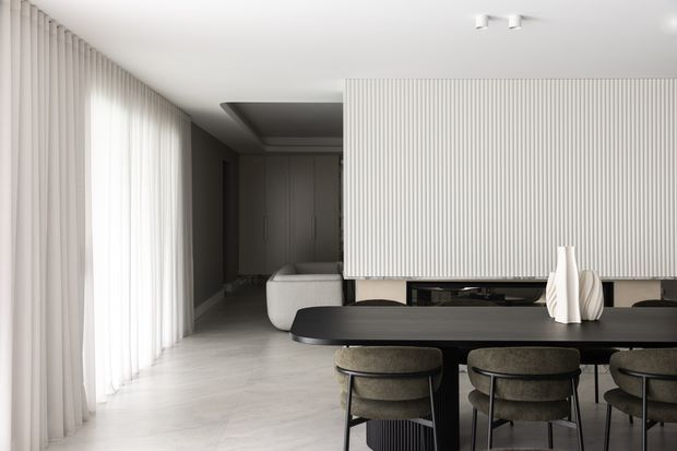 A refined contemporary dining area with a long black dining table and upholstered chairs in the foreground. Floor-to-ceiling sheer curtains line the left wall, while a large vertically ribbed feature screen or partition separates the dining area from a darker kitchen or living zone. The room uses a restrained palette of white, light grey, black, and muted taupe with clean built-in detailing and soft natural light.