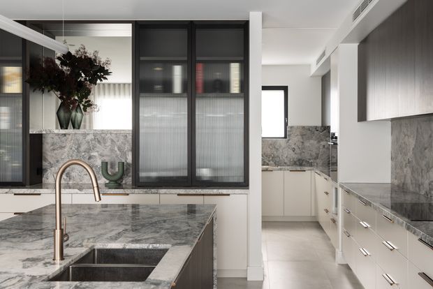 A refined contemporary kitchen with a large marble island, integrated double sink and brushed warm-metal tap. Pale cabinetry lines both sides of the room, while grey veined stone continues as the benchtops and splashback. Black-framed fluted-glass doors, a mirrored panel, and a floral arrangement add visual contrast. The space is bright, clean, and highly functional, with a secondary galley-style kitchen area visible beyond.