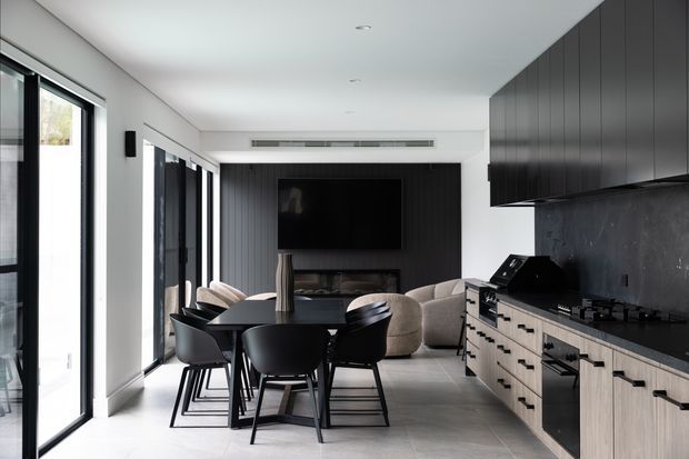 A bright, open-plan contemporary interior combining a kitchen, dining area and living room. The kitchen features dark upper cabinetry, light timber lower cabinetry, black benchtops and a dark splashback. A black dining table and chairs sit beside full-height glazing, while the living area includes a neutral sofa, dark feature wall, television and fireplace.