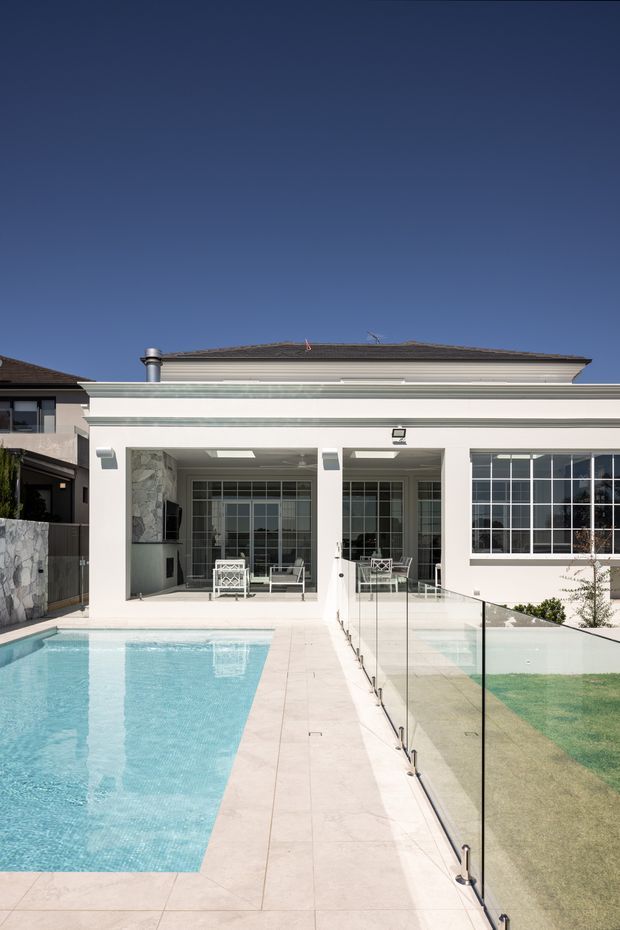 Exterior view of a refined residential home opening onto a swimming pool and paved terrace. The white rendered facade features a covered outdoor entertaining area, large multi-pane glazed doors and windows, a low hipped roof, and a frameless glass pool safety fence. The scene is bright and contemporary with pale paving and a turquoise pool.