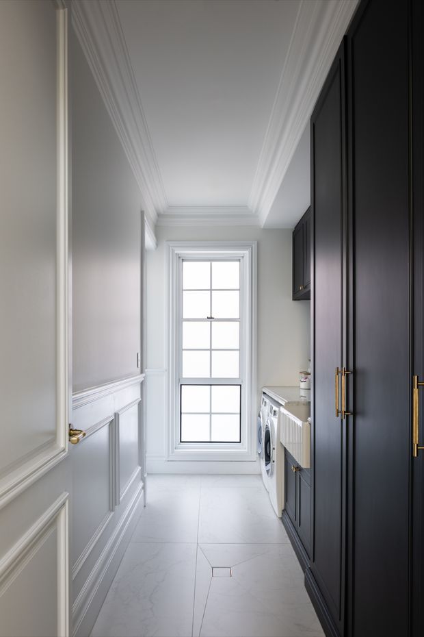 A narrow, elegant laundry room with a long galley layout. The room features dark charcoal-black full-height cabinetry with slim brass handles on the right, integrated laundry appliances and a light benchtop, while the left wall has pale grey decorative wainscoting and raised moulding. A tall multi-pane window provides natural light, and the space has large pale stone-look floor tiles and detailed white crown moulding.