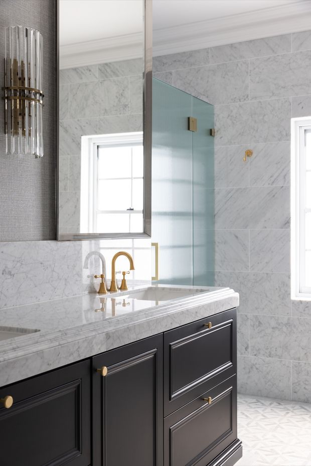 Elegant traditional bathroom vanity with black raised-panel cabinetry, white and grey veined marble wall tiles and benchtop, brass tapware and hardware, a framed mirror, and a fluted glass shower screen. Decorative moulding, gold accents, and patterned floor tiles create a refined classic look with bright natural light.