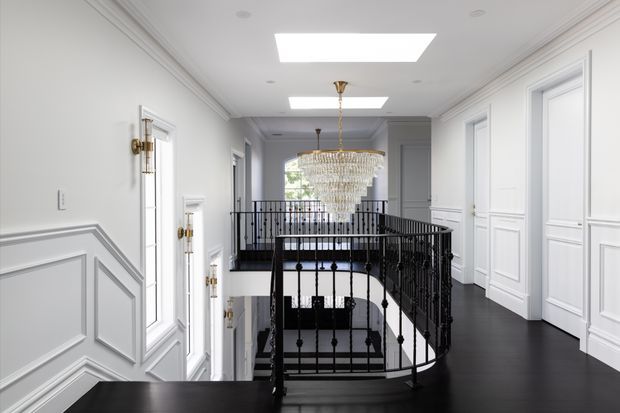 A bright, elegant upper-level landing and stair hall with a sweeping curved staircase, black metal balustrade, dark flooring, white decorative wall panelling and trim, multiple white paneled doors, and a large tiered crystal chandelier. Skylights and daylight create a refined, high-contrast monochromatic interior.