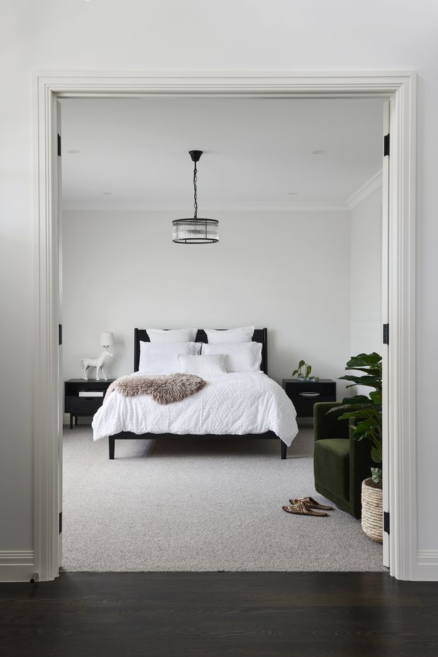 A bright, refined bedroom viewed through wide white-trimmed double doors. The room features a black upholstered bed with layered white bedding, black bedside tables, a pendant light, pale carpet, green armchair, indoor plants, and dark timber flooring in the foreground. The overall look is clean and contemporary with monochrome furnishings and soft natural accents.