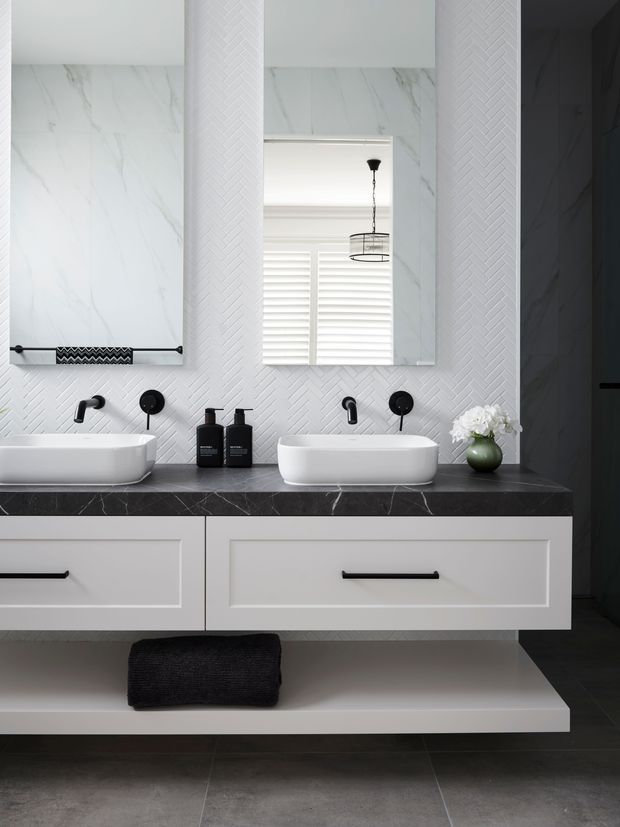 A refined contemporary bathroom vanity with two white vessel basins on a dark marble-look benchtop. The wall is clad in white herringbone-pattern tile, complemented by large frameless mirrors, matte-black tapware and handles, white drawers, and a warm dark grey tiled floor. A small green vase with white flowers and black towels add restrained accents.