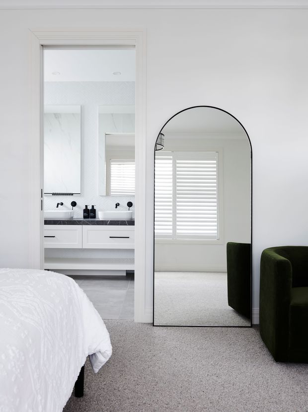 Bright, refined bedroom with a white upholstered bed partially visible in the foreground, light grey carpet, and a dark green upholstered armchair. An arched black-framed full-length mirror reflects a shuttered window and carpeted room. Through an open doorway, a white double vanity bathroom is visible, featuring black tapware, dark stone benchtops, white cabinetry, and pale marble-look wall surfaces.
