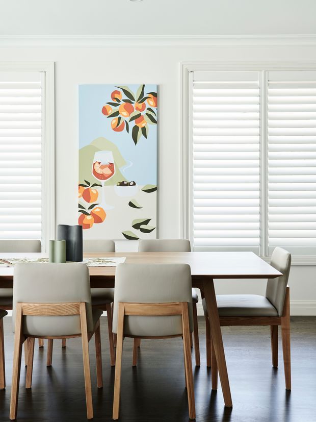 Bright dining room with a rectangular light-coloured dining table and upholstered wooden dining chairs. White plantation shutters cover the windows, while a colourful botanical-style artwork provides a focal point. The room has light walls, dark timber flooring, and a clean, contemporary look.