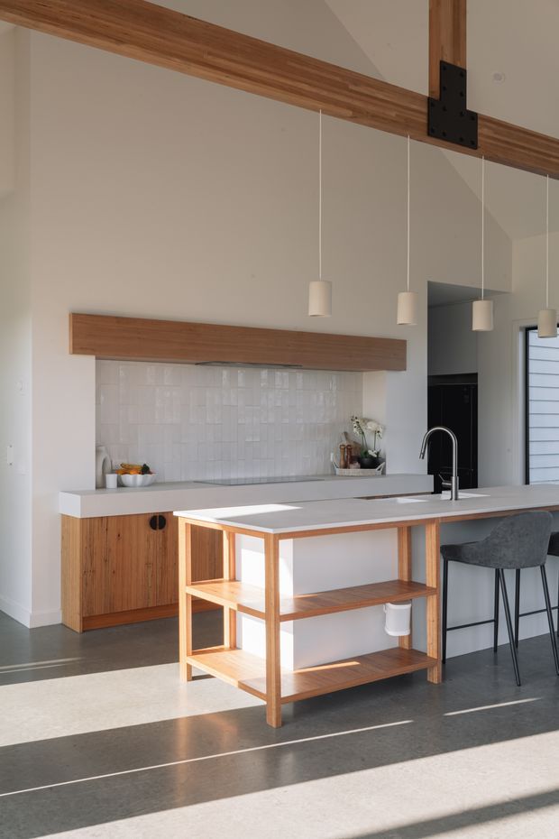 Bright contemporary kitchen with a large island featuring open timber shelving, a white benchtop, integrated sink and tall curved tap. The kitchen includes warm timber cabinetry and overhead timber beam detailing, a glossy white textured tile splashback, minimalist pendant lights, dark flooring, and bar seating. Strong sunlight creates long shadows across the floor.