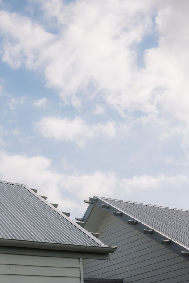 Upward-looking exterior architectural detail showing overlapping pitched gable roofs with light grey corrugated metal roofing, pale horizontal weatherboard cladding, exposed roof edges and gutters, against a partly cloudy blue sky.