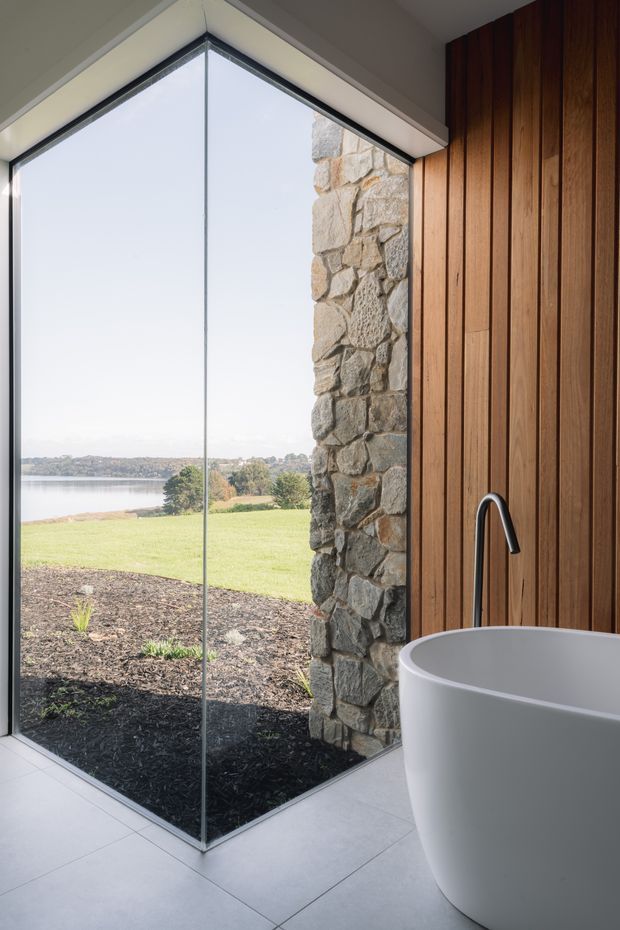 A contemporary bathroom corner with a freestanding white bathtub and sleek black tapware beside floor-to-ceiling corner glazing. The space features a vertical timber-lined wall, a rugged natural-stone feature wall, and large pale floor tiles. Through the glazing is a landscaped lawn and visible body of water, creating a bright, open outlook.
