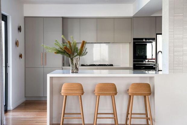 A bright, contemporary kitchen with a large white island and three timber bar stools. The kitchen features full-height matte grey cabinetry, integrated appliances, a pale stone-look benchtop and splashback, warm timber flooring, and minimalist white walls. A vase of greenery and dried stems provides a natural decorative accent.