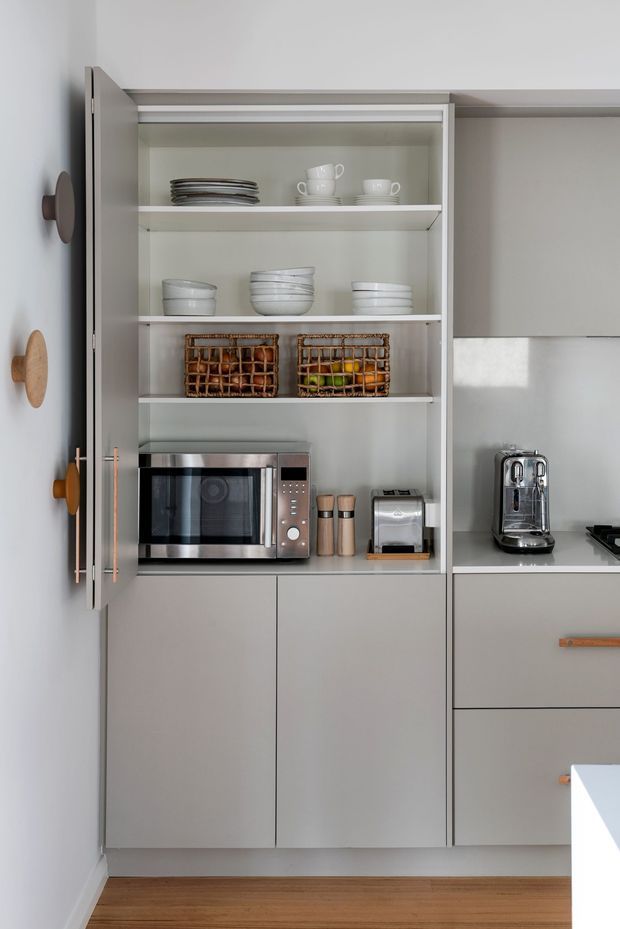 A compact, contemporary kitchen focused on a tall built-in pantry and appliance cabinet. The cabinetry has smooth light-grey doors and open shelving displaying crockery and woven baskets, with integrated microwave, toaster, coffee machine and countertop appliances. A pale stone or composite benchtop, white splashback, and warm timber flooring create a clean, restrained interior.