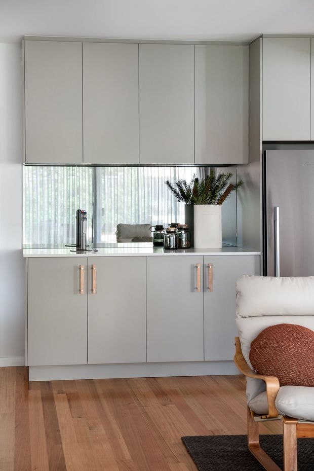 A compact, contemporary open-plan kitchen and living area with full-height pale grey cabinetry, integrated refrigerator, light stone benchtop, and vertical white sheer blinds over a large window. Warm timber flooring contrasts with the cool cabinetry, while a white lounge chair with a rust-coloured cushion occupies the foreground. A vase of greenery and small kitchen accessories add subtle decoration.