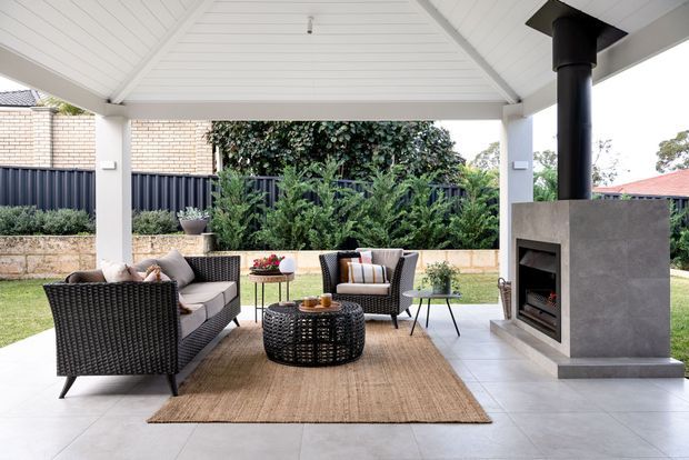 Covered outdoor entertaining area with a white-lined pitched ceiling, tiled floor, woven outdoor lounge furniture, a jute rug, and a substantial grey rendered fireplace with black flue. The space opens onto a landscaped garden and lawn, with a masonry boundary wall and dark fencing beyond.