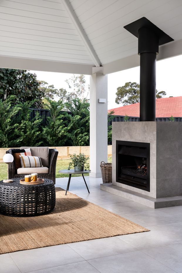 A covered outdoor entertaining area with a built-in dark rendered fireplace and black flue beneath a white-lined pitched roof. The space includes woven outdoor lounge chairs, a round woven coffee table, a small side table, a natural-fibre rug, tiled flooring, and garden views beyond the open sides.