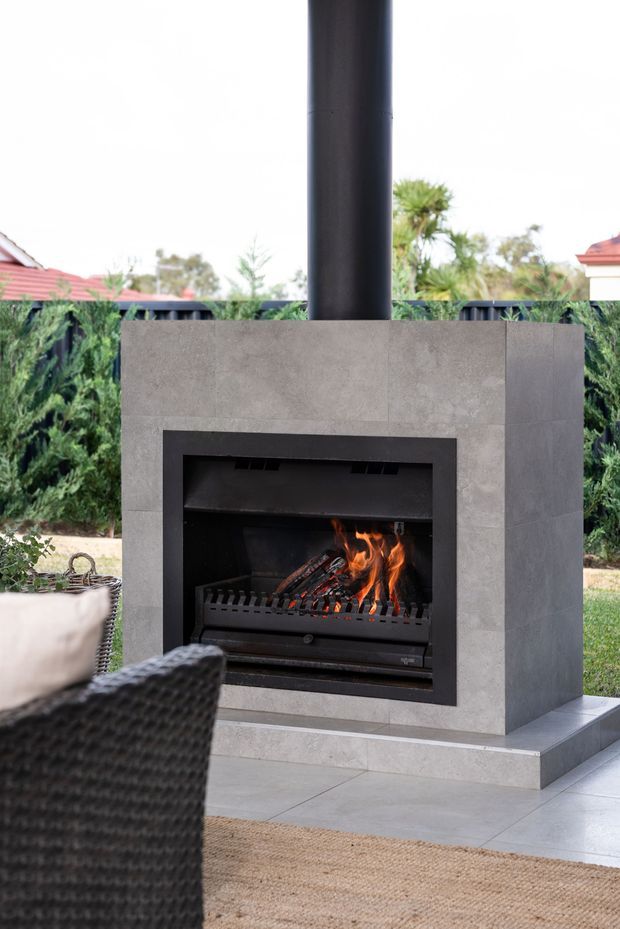 A contemporary outdoor fireplace clad in large grey stone or concrete-look panels, with a black metal firebox, chimney flue, and visible flames. It sits on a tiled patio beside woven outdoor seating, with landscaped greenery and neighbouring rooflines in the background.