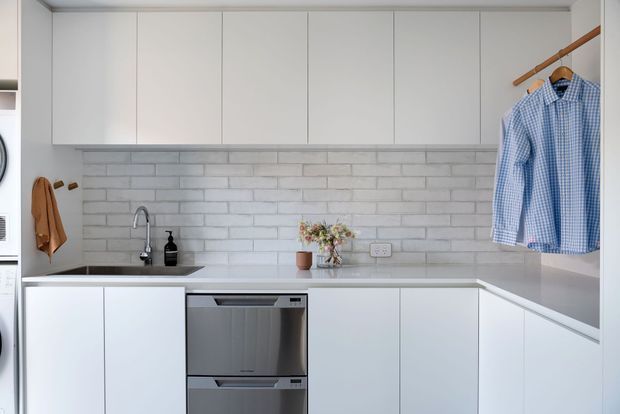Bright, compact contemporary laundry with white handleless cabinetry, a pale grey-white brick-look tiled splashback, stainless-steel dishwasher, integrated sink and tap, wall-mounted hanging rail, and a small benchtop arrangement. The layout wraps around two walls and uses a clean, minimal palette.