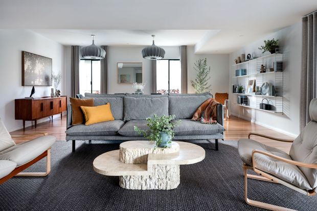Bright, refined living room with a large grey sofa, sculptural round coffee tables, lounge chairs, timber mid-century-style sideboard, open wall shelving, pendant lights, and floor-to-ceiling curtains. The room combines contemporary finishes with warm natural timber, textured textiles, and organic stone-like furniture forms.