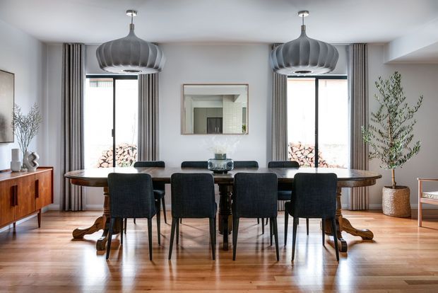 A refined dining room with a long dark timber dining table surrounded by upholstered black dining chairs. The room features polished timber flooring, white walls and ceiling, tall windows with striped floor-length curtains, sculptural pendant lights, a sideboard, and restrained decorative greenery.