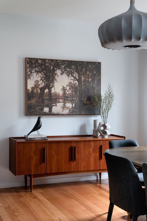 A warmly styled dining room with a mid-century-inspired timber sideboard, dark upholstered dining chairs, and a timber dining table. Pale walls and timber flooring create a calm neutral backdrop, while a large framed landscape artwork, sculptural pendant light, ceramic vase, branches, and decorative objects add character.